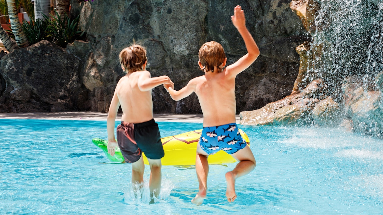 Kids jumping into pool at water park with waterfall and inflatable float.