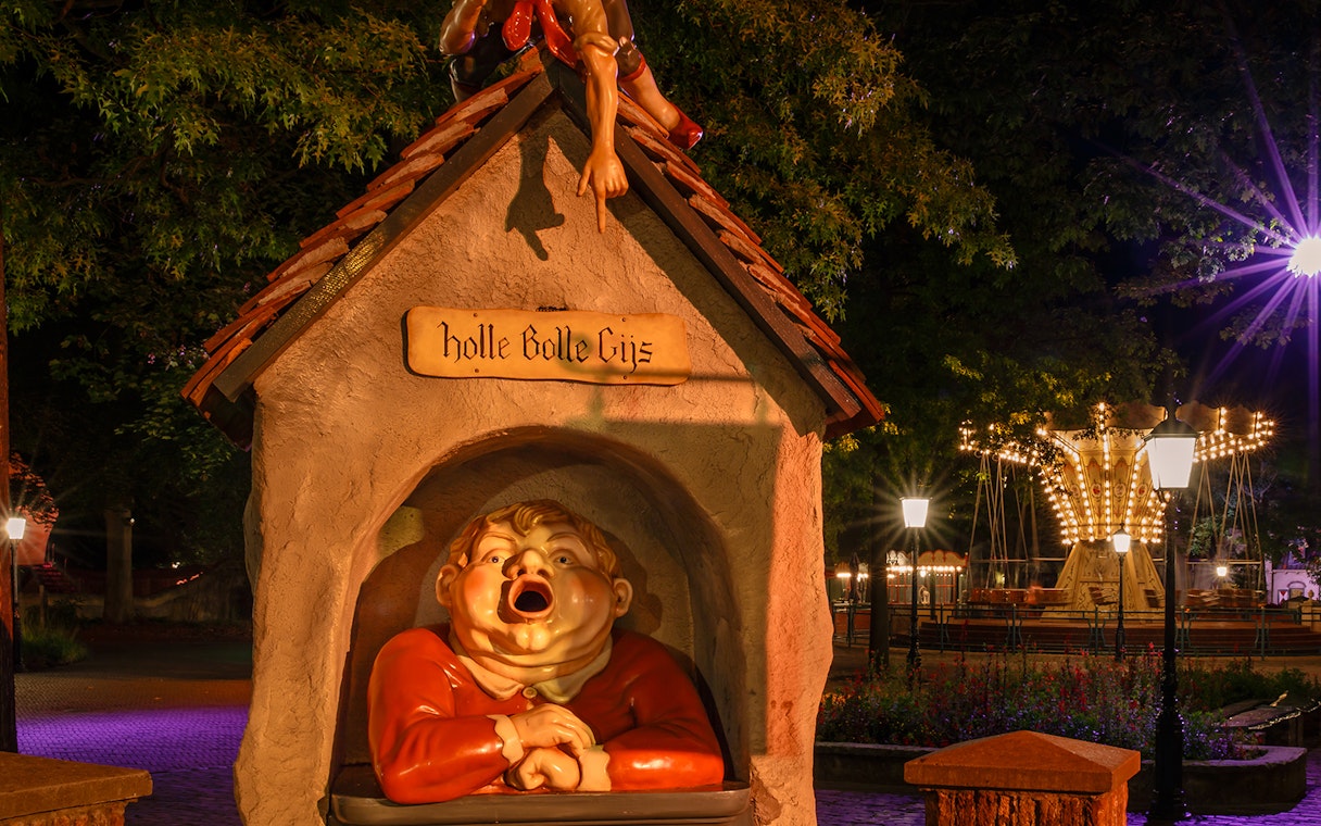 Holle Bolle Gijs statue at Efteling theme park, Netherlands, with illuminated carousel in background.