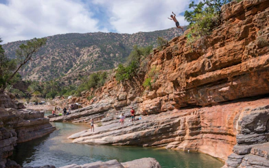 Cliff diver at Paradise Valley, Morocco, with rocky landscape and people by the water.
