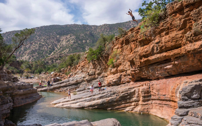 Cliff diver at Paradise Valley, Morocco, with rocky landscape and people by the water.