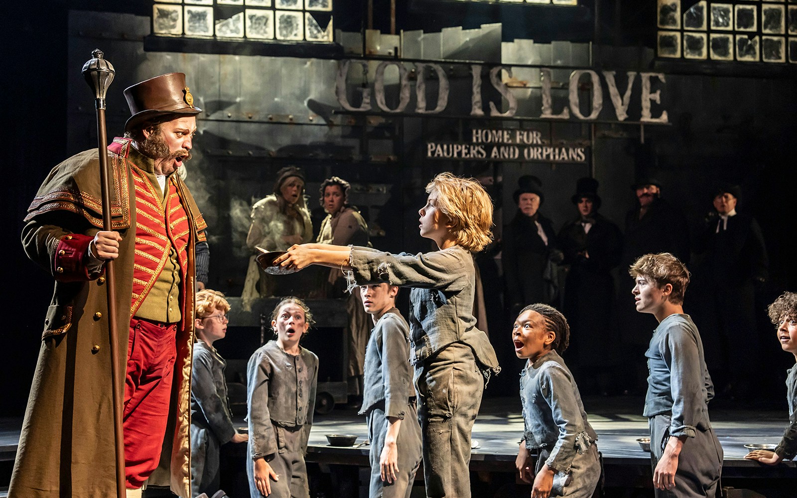 Child holding a bowl towards a man in a theatrical scene from Oliver!