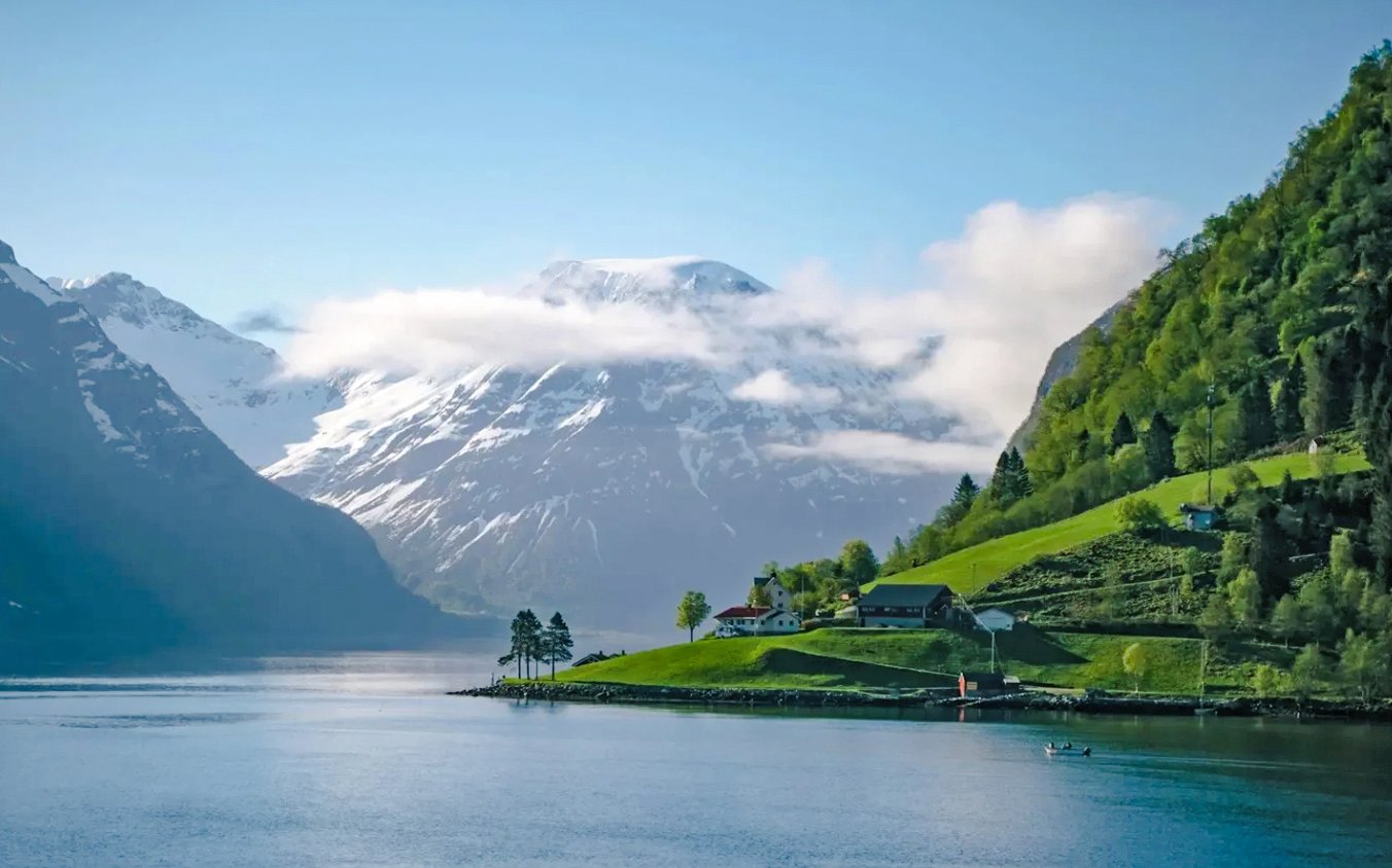 Houses on a green hillside by Hjørundfjord, Norway with snow-capped mountains in the background.