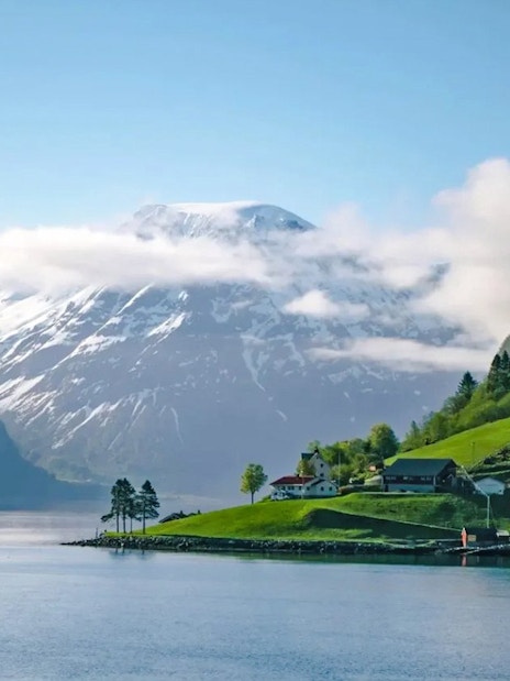 Houses on a green hillside by Hjørundfjord, Norway with snow-capped mountains in the background.
