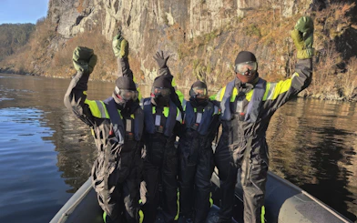 Passengers in safety suits celebrating on a fjord boat with rocky cliffs in the background.