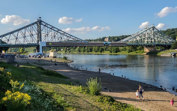 Steel bridge over the Elbe River in Dresden with people walking along the riverbank.