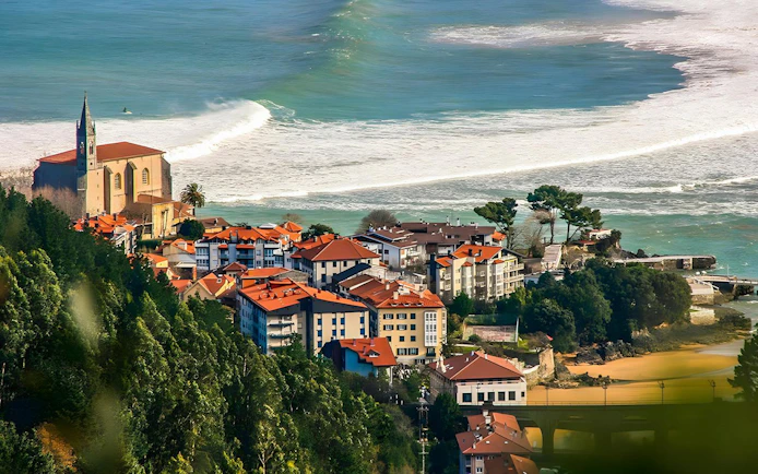 Arial view of Mundaka, Spain, showing coastal village with red-roofed buildings and church by the ocean.
