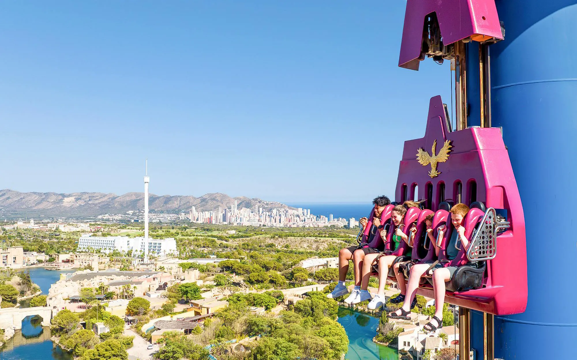 Visitors on a drop tower ride at Terra Mitica theme park, Spain, with cityscape in the background.