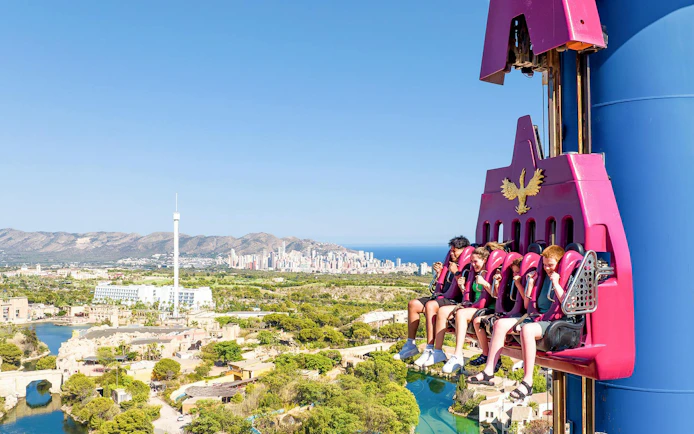 Visitors on a drop tower ride at Terra Mitica theme park, Spain, with cityscape in the background.