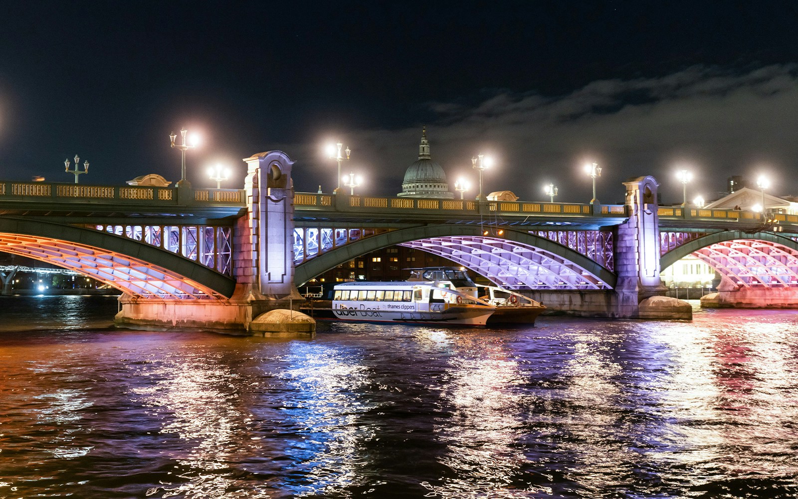 Uber Boat on Thames River passing under illuminated London bridge at night.