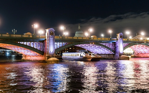 Uber Boat on Thames River at night passing under illuminated London bridge.