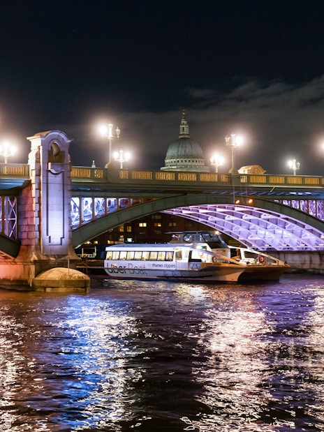 Uber Boat on Thames River at night passing under illuminated London bridge.