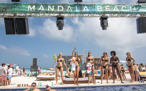People enjoying a sunny day at Mandala Beachclub Cancun, with a pool and beach in the background.