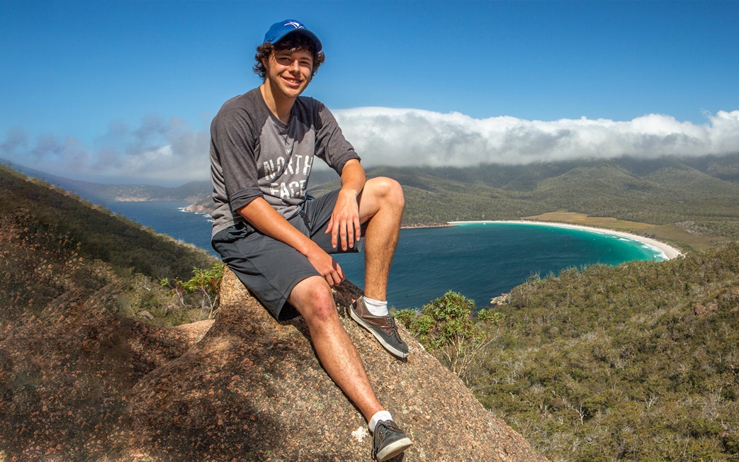 Person sitting on a rock overlooking Wineglass Bay, Tasmania, with lush hills and blue water.