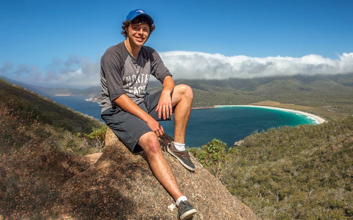 Person sitting on a rock overlooking Wineglass Bay, Tasmania, with lush hills and blue water.