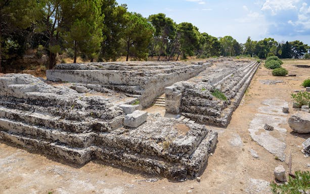 Ancient stone ruins at Neapolis Archaeological Park, Syracuse, surrounded by trees.