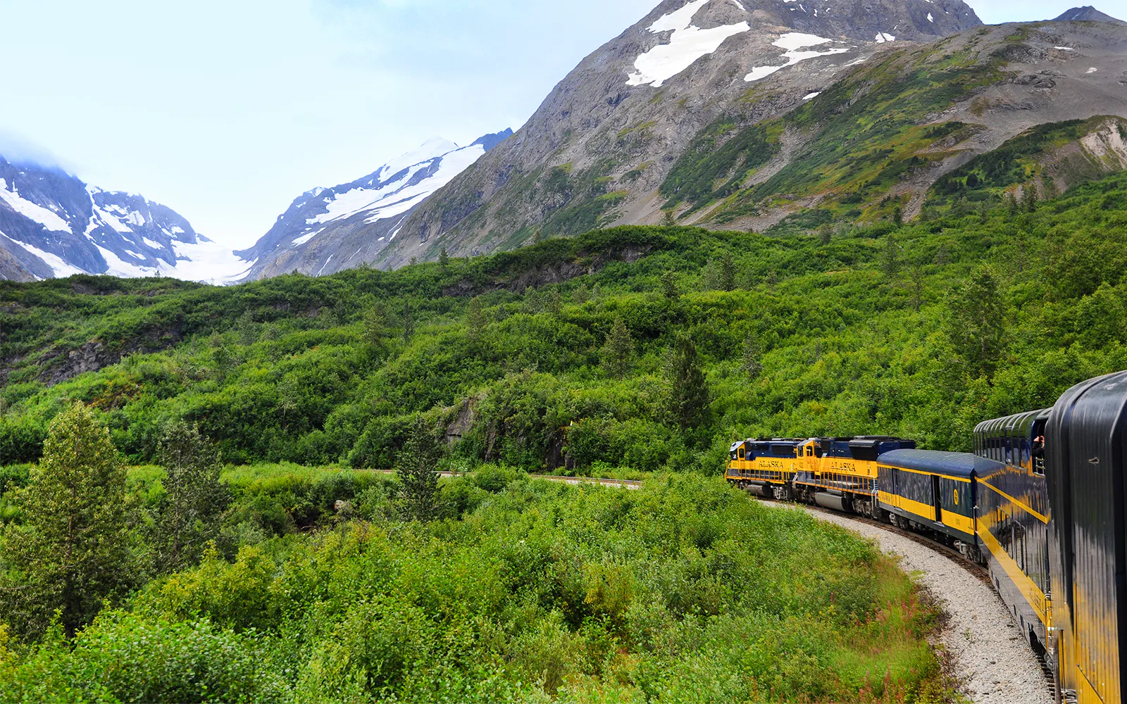 Alaska Railroad train traveling through lush green mountains with snow-capped peaks.