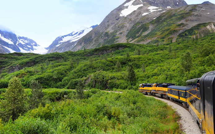 Alaska Railroad train traveling through lush green mountains with snow-capped peaks.