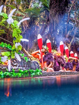 Mitai Maori Village performers paddling a traditional canoe in a lush forest setting.