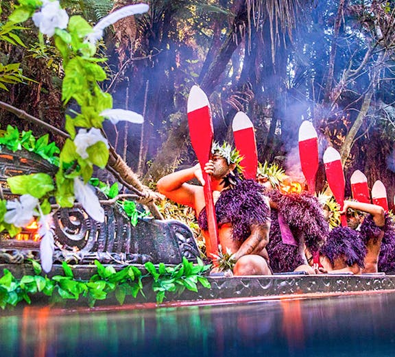 Mitai Maori Village performers paddling a traditional canoe in a lush forest setting.