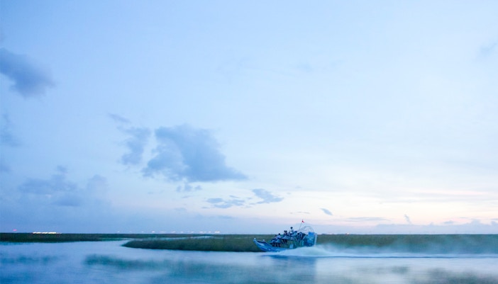 An airboat glides across the tranquil waters of the Florida Everglades at dusk, its wake creating a subtle mist trail behind it. The soft blue of the sky and water, punctuated by distant greenery, evokes a sense of adventure and peace.