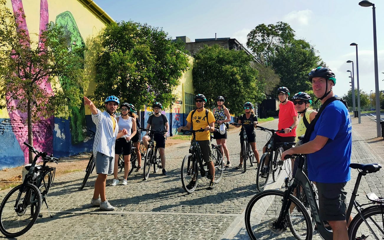 Group on electric bikes touring Athens with guide pointing at street art.