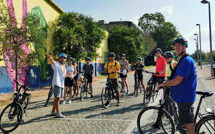 Group on electric bikes touring Athens with guide pointing at street art.