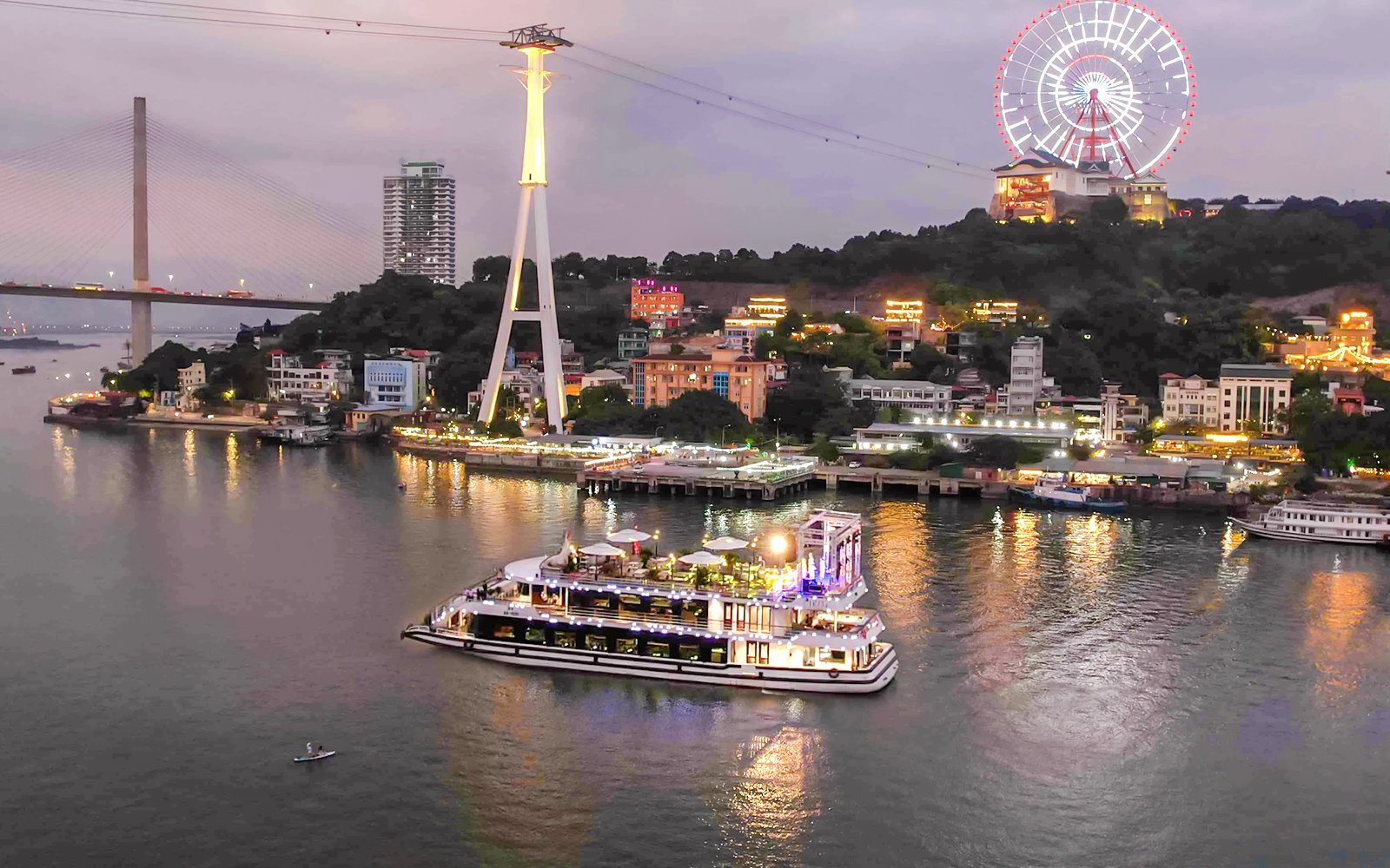 Iris Luxury Cruise sailing in Ha Long Bay during evening with city lights and Ferris wheel.
