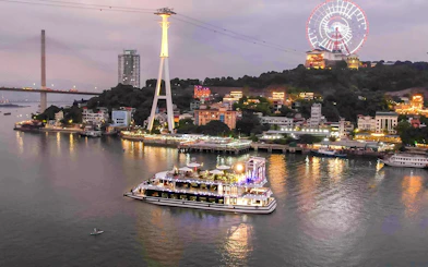 Iris Luxury Cruise sailing in Ha Long Bay during evening with city lights and Ferris wheel.