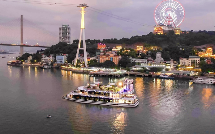 Iris Luxury Cruise sailing in Ha Long Bay during evening with city lights and Ferris wheel.