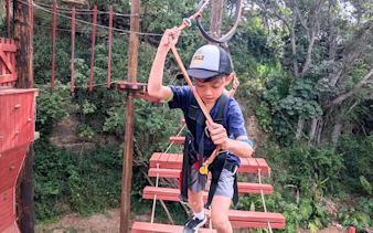 Child navigating high-elevation adventure course with safety gear in forest setting.