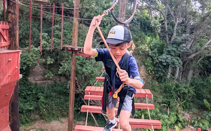 Child navigating high-elevation adventure course with safety gear in forest setting.