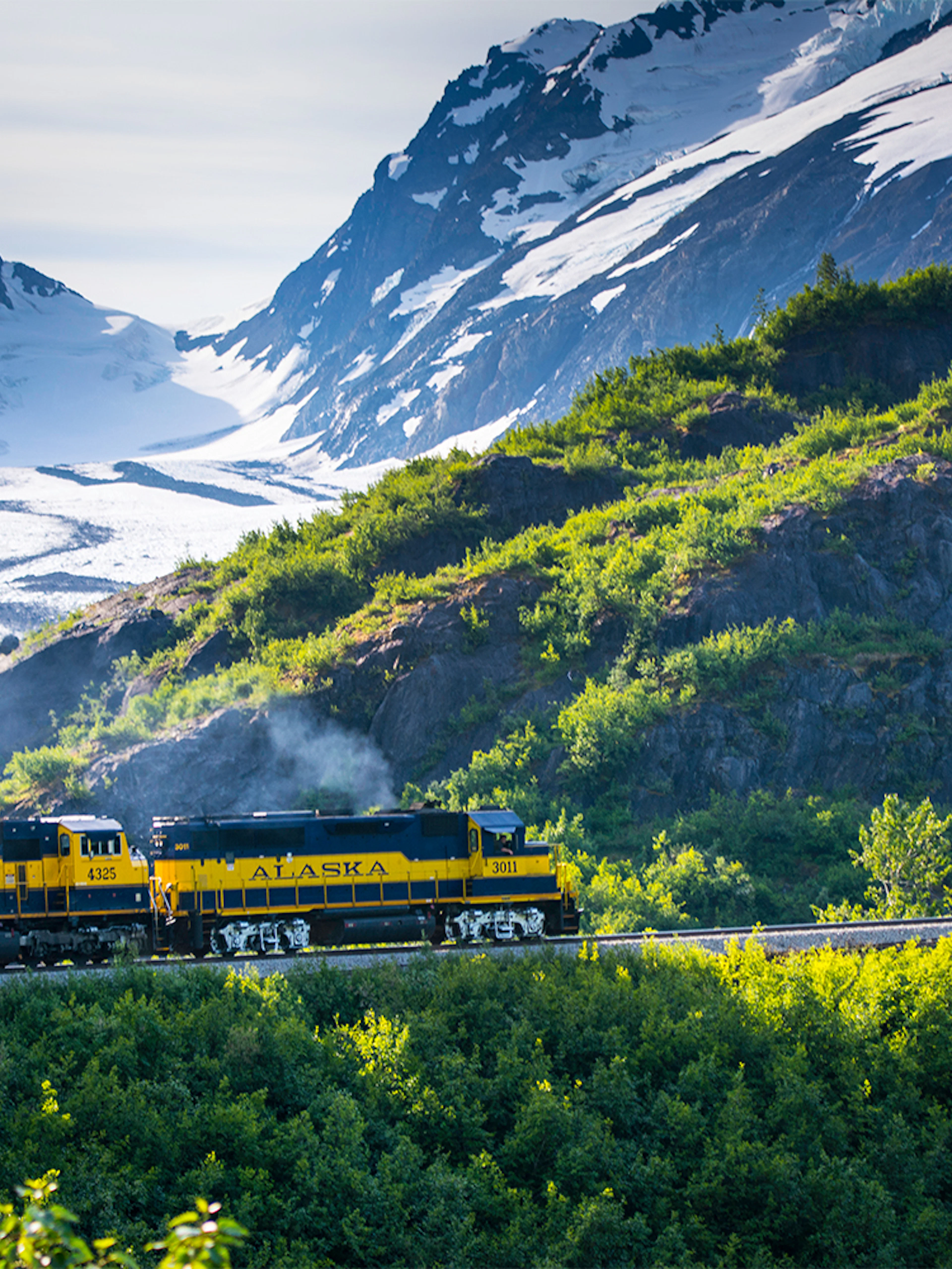 Alaska Railroad train traveling through lush green mountains with snow-capped peaks in the background.