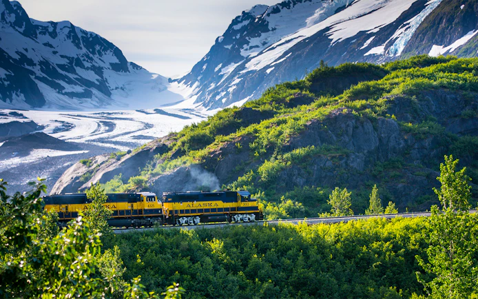 Alaska Railroad train traveling through lush green mountains with snow-capped peaks in the background.
