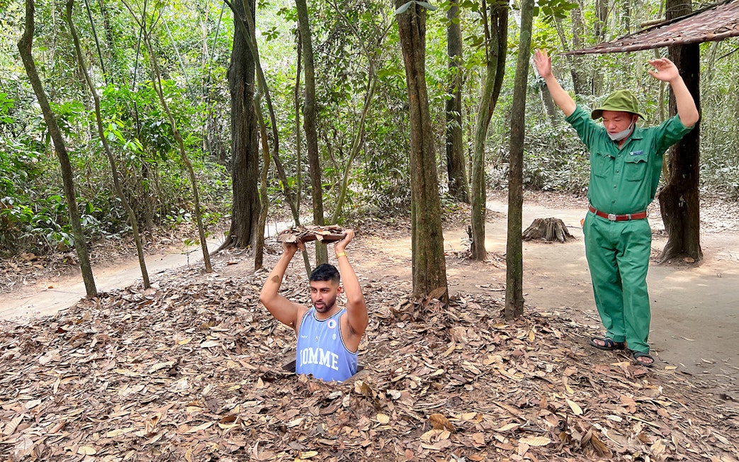 Man emerging from Cu Chi Tunnel holding cover, with guide nearby in forest setting.