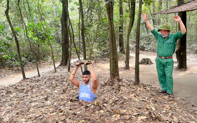 Man emerging from Cu Chi Tunnel holding cover, with guide nearby in forest setting.