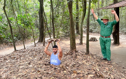 Man emerging from Cu Chi Tunnel holding cover, with guide nearby in forest setting.
