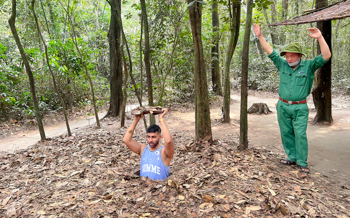 Man emerging from Cu Chi Tunnel holding cover, with guide nearby in forest setting.