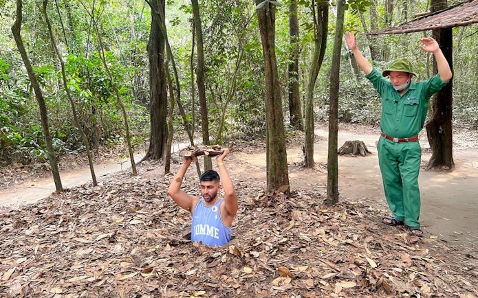 Man emerging from Cu Chi Tunnel holding cover, with guide nearby in forest setting.