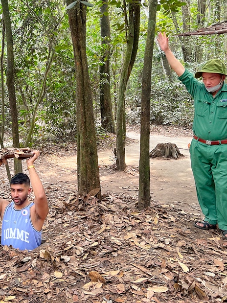 Man emerging from Cu Chi Tunnel holding cover, with guide nearby in forest setting.