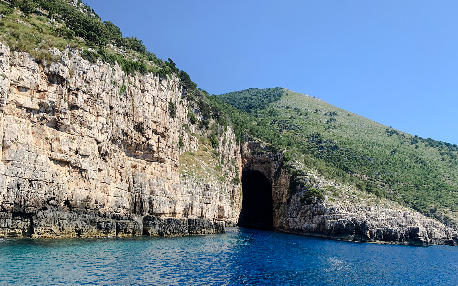 Haxhi Ali Cave entrance along rocky coastline with clear blue water in Albania.