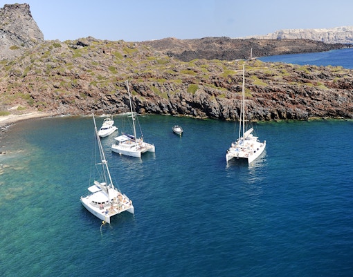 Catamarans sailing near rocky coast on Santorini luxury cruise.
