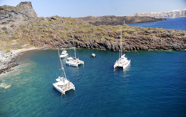 Catamarans sailing near rocky coast on Santorini luxury cruise.