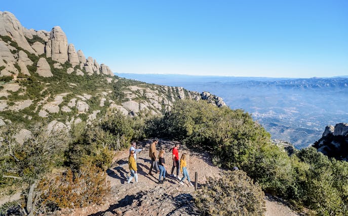 Tourists hiking with guide on trail to Montserrat Monastery, Spain, with mountain views.