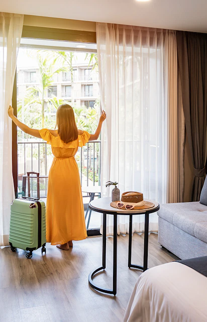 Person in a hotel room with luggage, looking out at a garden view.