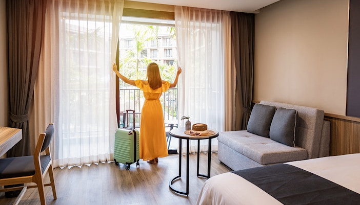 Person in a hotel room with luggage, looking out at a garden view.