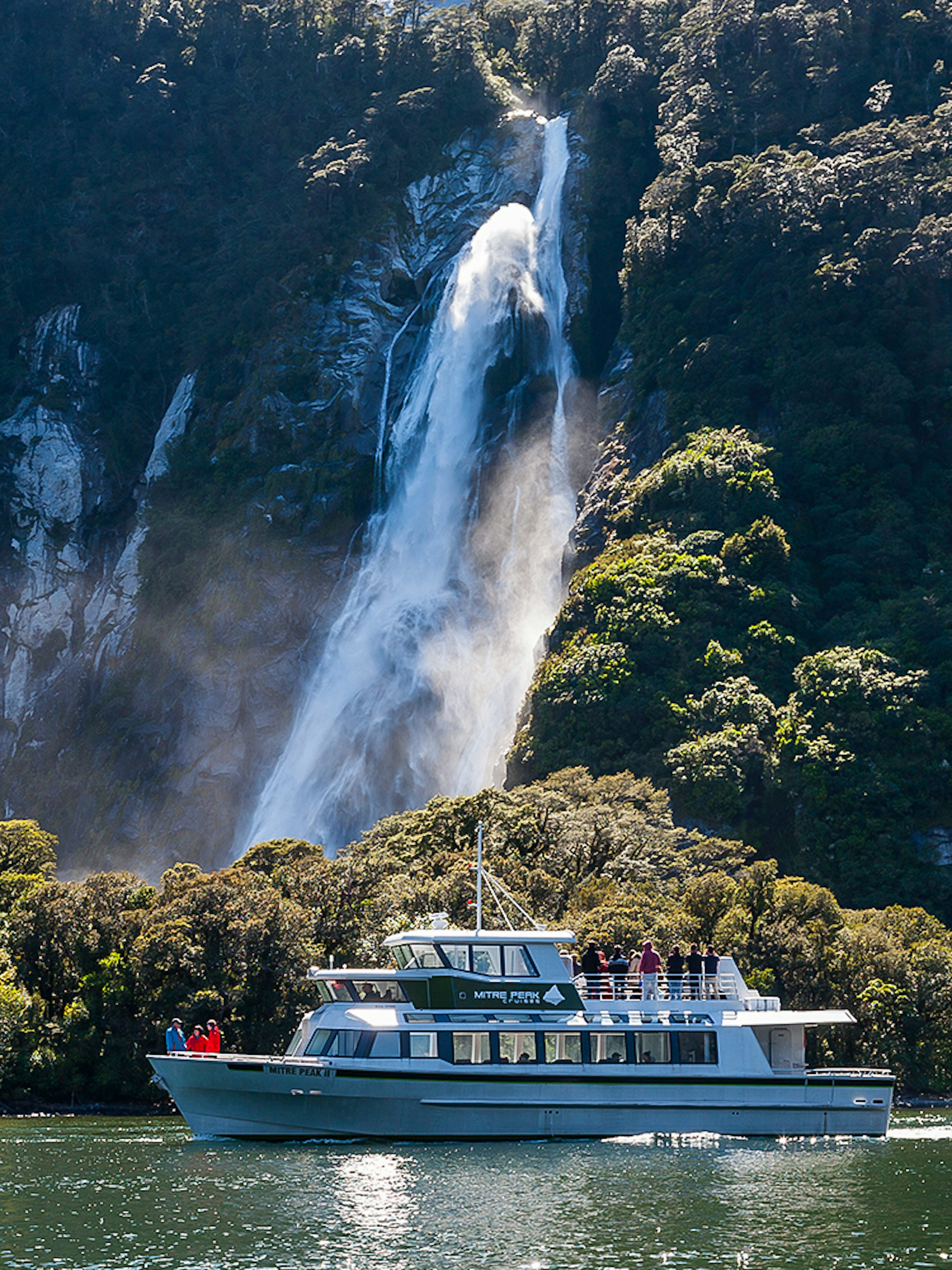 Small boat cruising near waterfall in Milford Sound, New Zealand.