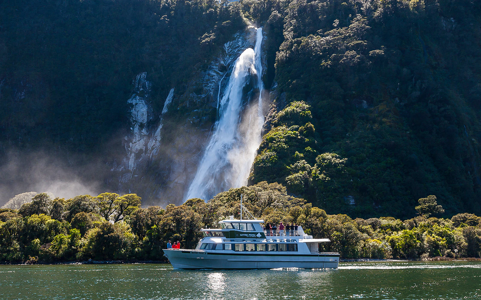Small boat cruising near waterfall in Milford Sound, New Zealand.