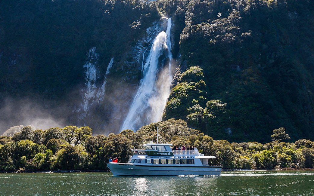 Small boat cruising near waterfall in Milford Sound, New Zealand.