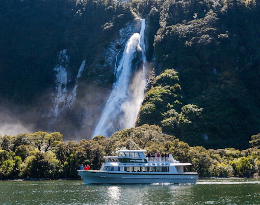 Small boat cruising near waterfall in Milford Sound, New Zealand.
