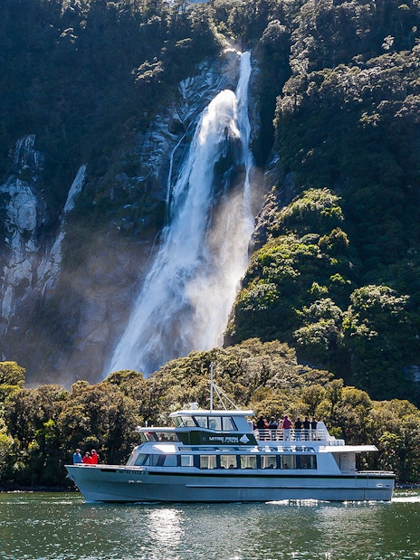 Small boat cruising near waterfall in Milford Sound, New Zealand.